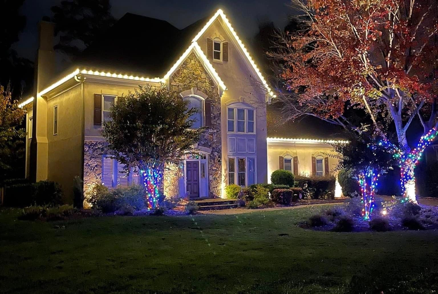 Home with warm white roofline lights and color-wrapped trees at night